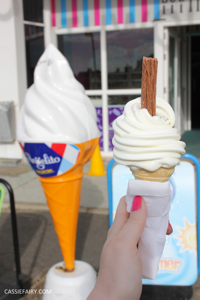 A hand holding an ice cream cone with a flake, beside a large plastic ice cream sculpture, outside a sweet shop.