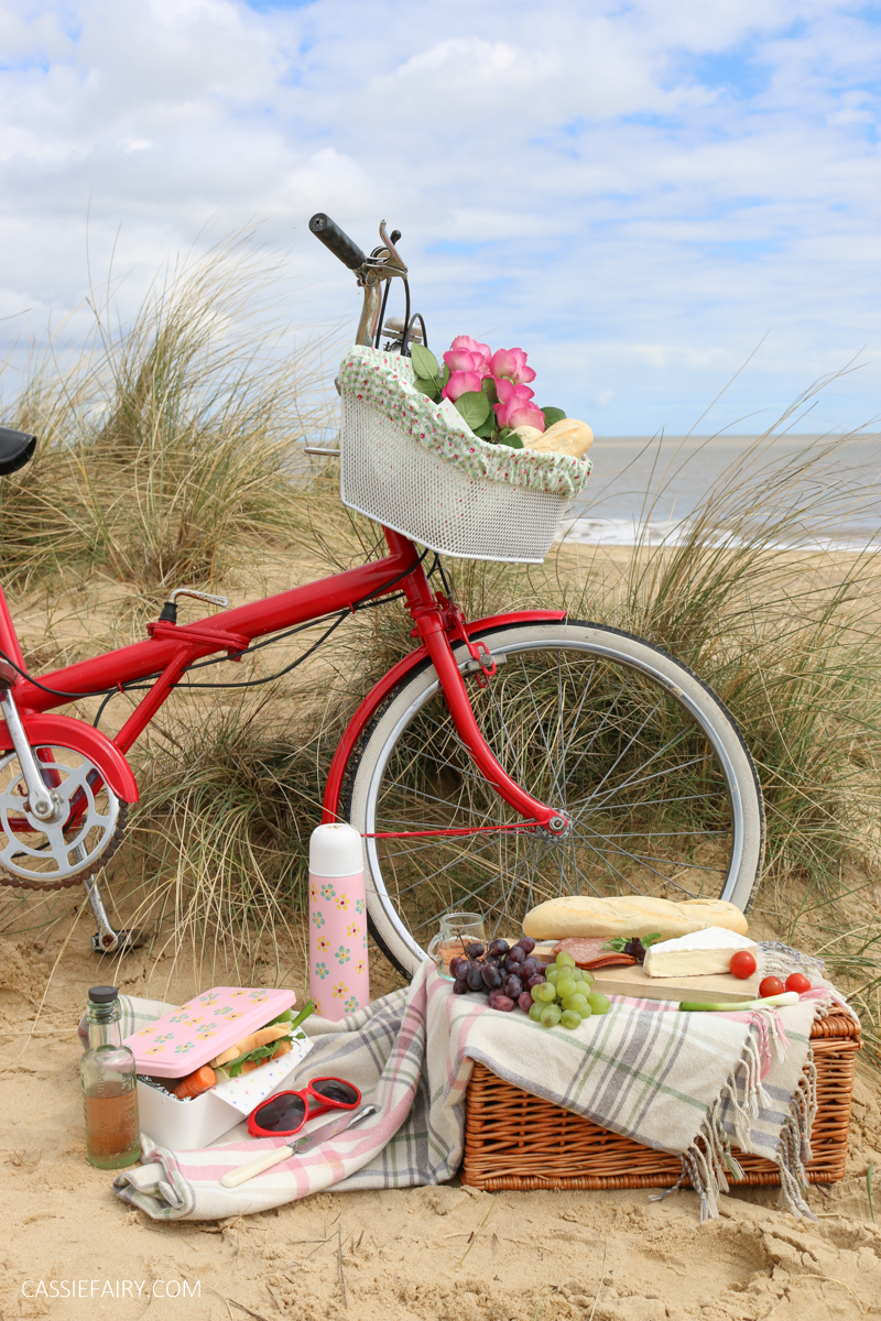 The dunes of a sandy beach with a food arranged on a picnic hamper in front of a red bicycle.