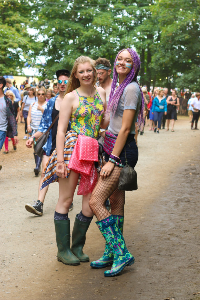 Two girls posing at a music festival wearing denim shorts, colourful printed tops and wellies. One girl has purple plaited hair.