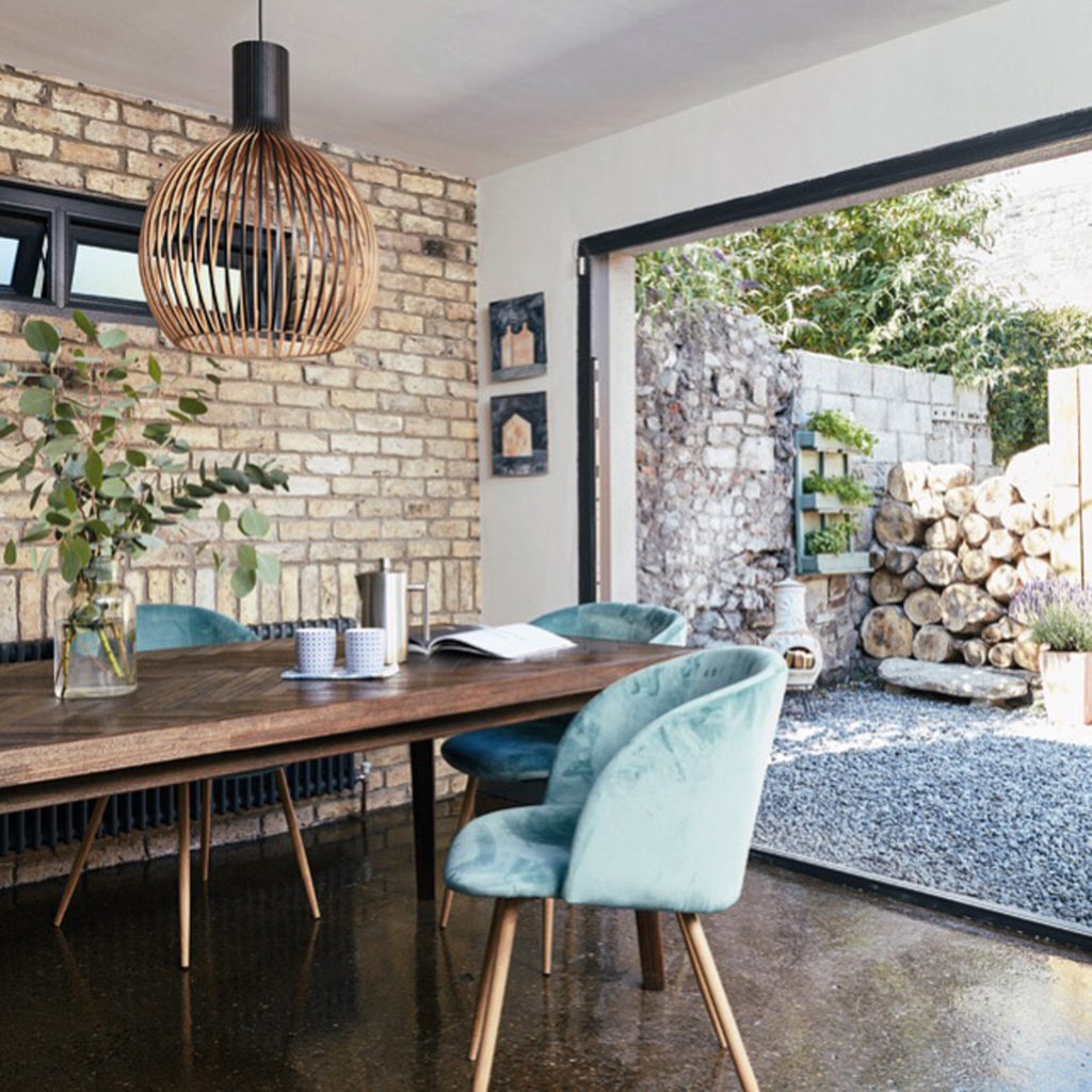 An extension with a large patio window and an exposed brick wall. Inside the room is a wooden dining table surrounded by pale turquoise velvet dining chairs. 