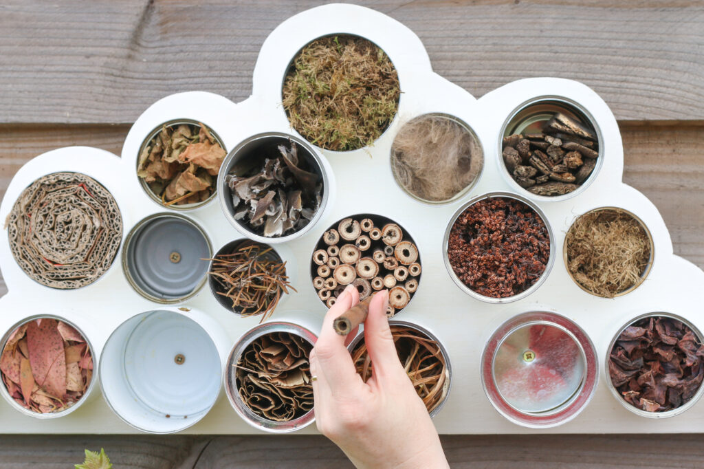 Image shows cuttings of dried seed heads, leaves, grasses, bark, moss and wool filling the tin cans on the bug hotel