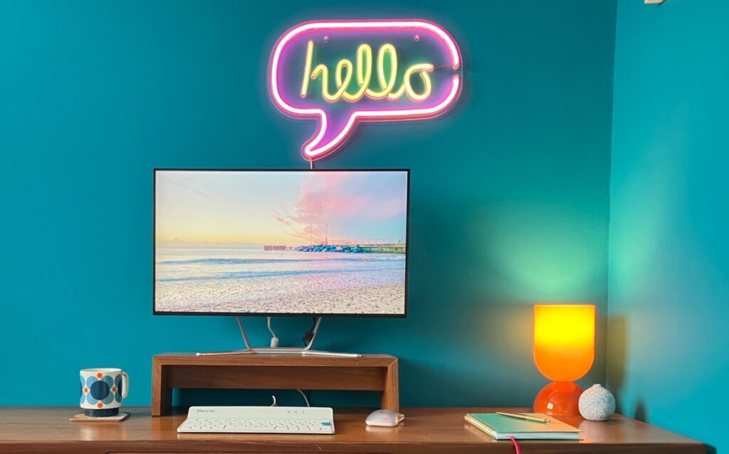 A home office desk with a monitor, keyboard and an orange lamp, with a neon sign on the turquoise wall that shows the word 'Hello' lit up in pink neon.   