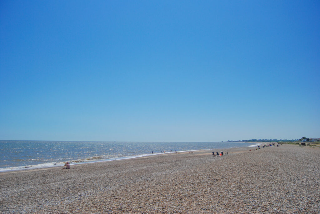 A bright blue sky with no clouds above a shingle beach with the sea rolling into the shore on the left.