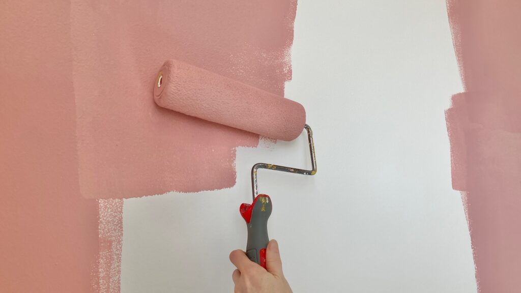 A hand holding a roller, painting pink paint onto a white wall.