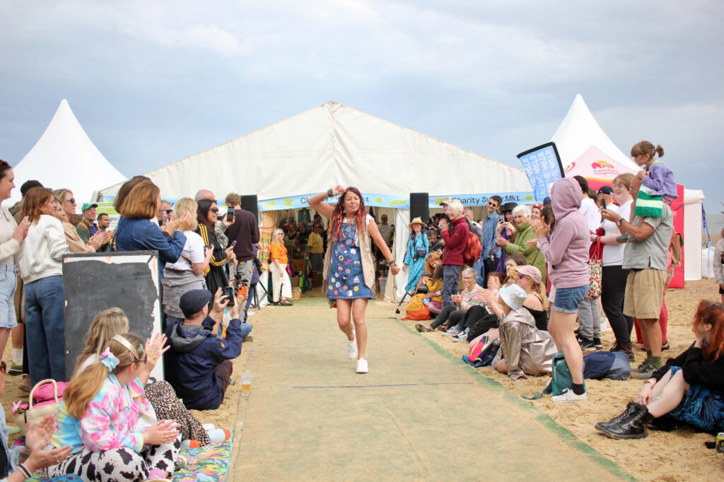 A red-haired model on a catwalk at a festival with an audience at either side, wearing a customised denim dungaree dress covered in patches, along with a fringed beige waistcoat and white trainers.