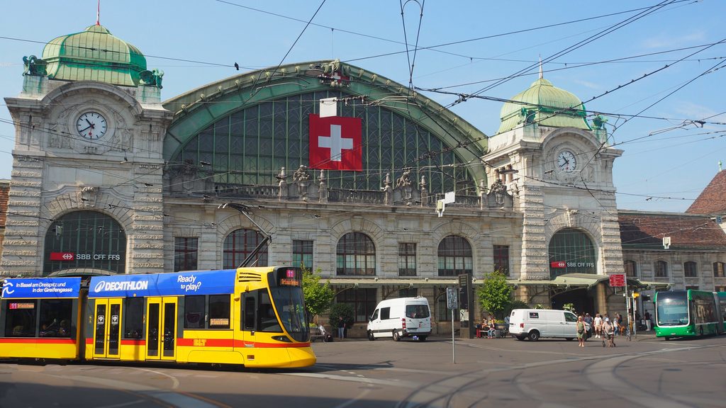 The exterior of a train station in Switzerland with a tram driving past it.