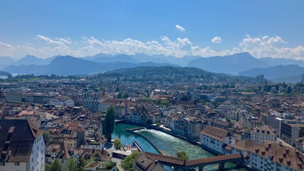 Landscape image of Lucerne in Switzerland, taken from a high tower, showing the river with buildings either side of it and the mountains on the horizon and clouds in the sky.