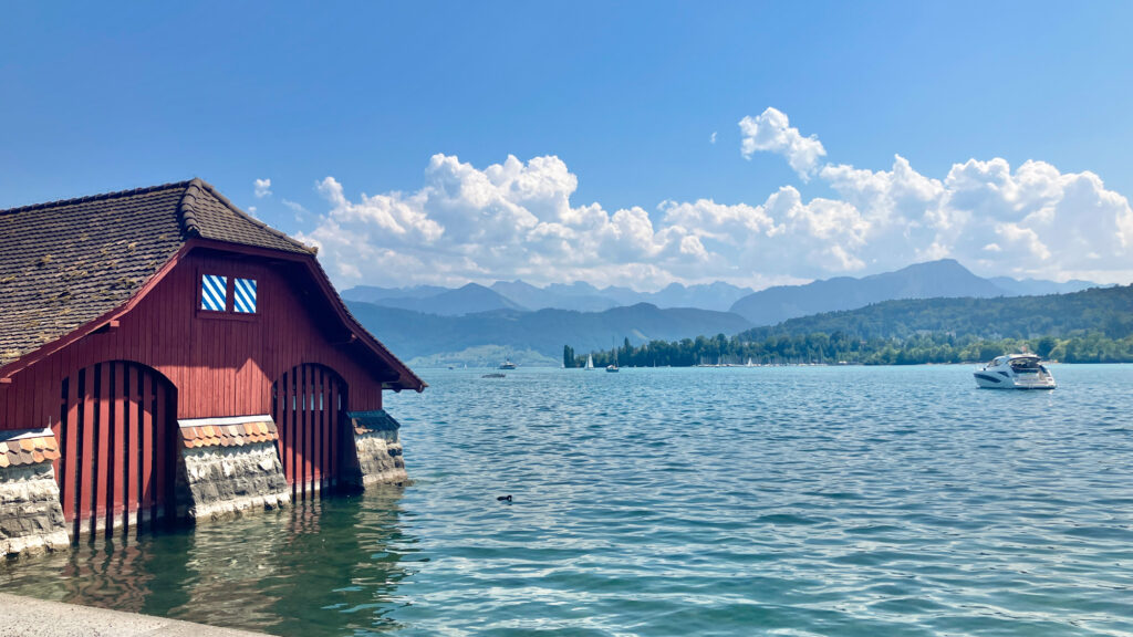 A view over the blue waters of Lake Lucerne with the mountains on the horizon and clouds in the blue sky. In the foreground is a red-painted boathouse on the shore of the lake. 
