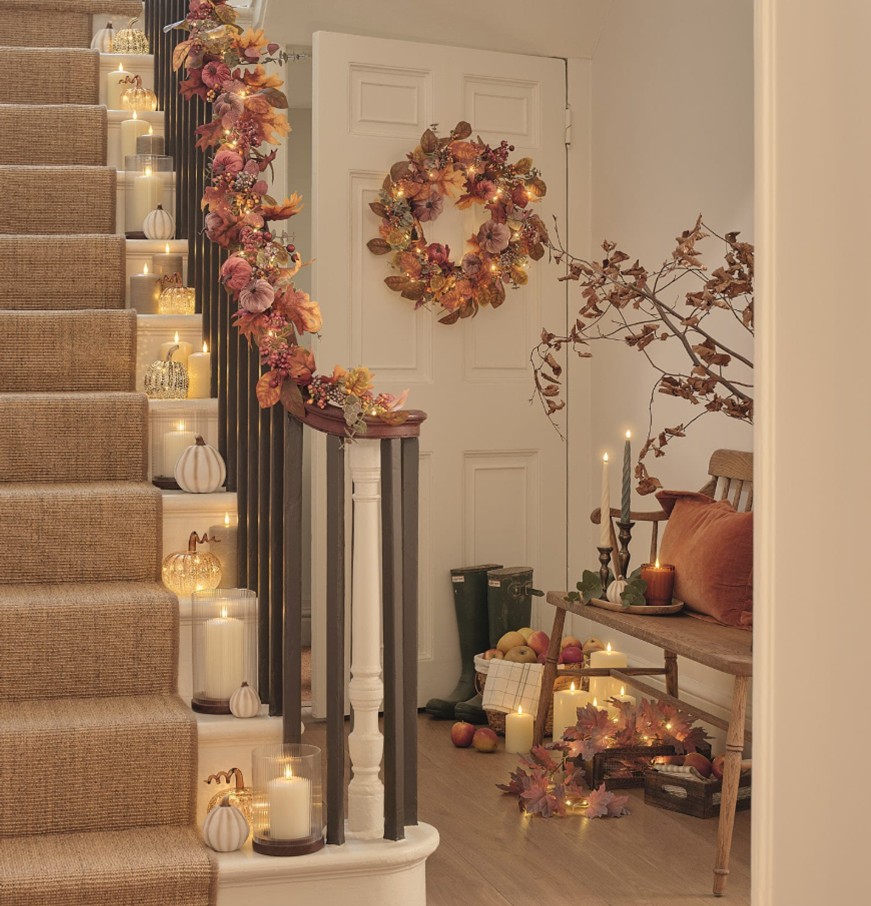 A white hallway and a staircase with a jute runner, decorated with faux autumnal foliage garlands, fairy lights and LED candles on each step.