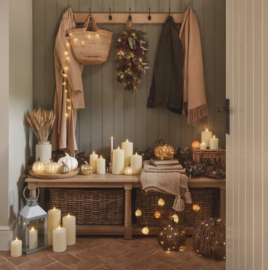 An entryway with a wooden bench and coat hooks on the painted shiplap wall, decorated with fairy lights, LED candles, blankets and baskets.