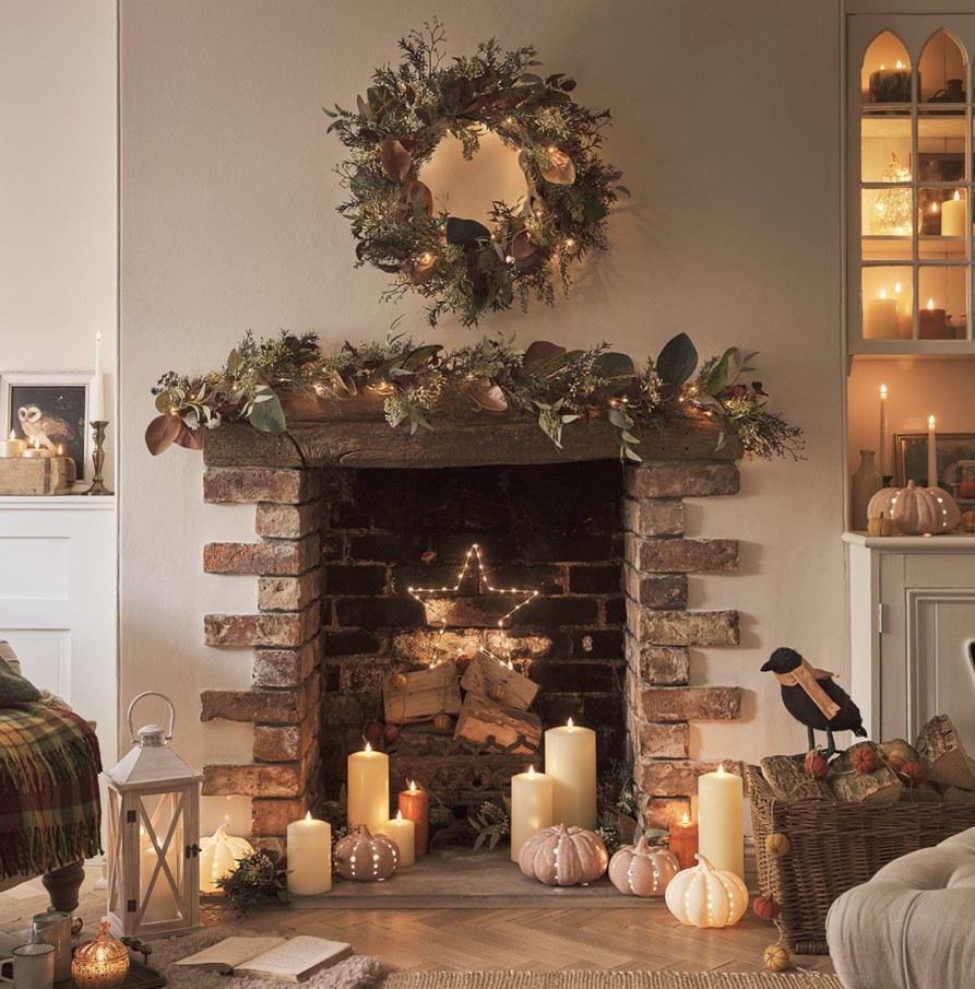 A brick fireplace with a wooden mantlepiece, decorated with a faux autumn garland and a pre-lit wreath, with LED candles and pumpkins on the hearth.