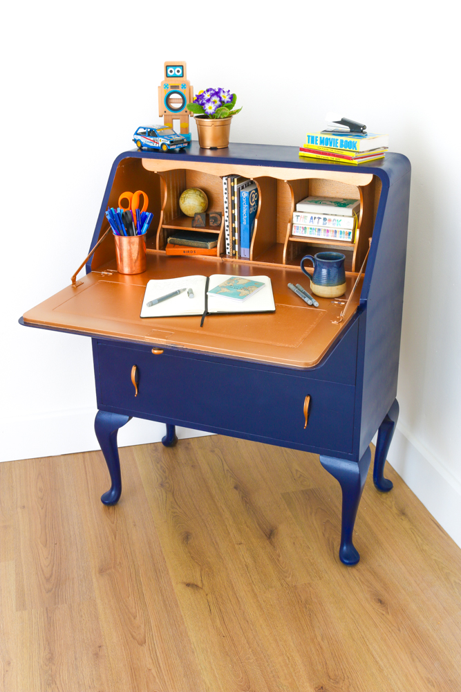 The finished dark blue bureau with its desk open with a a copper interior, styled with a notebook, books, a copper pot of pens and blue cup of coffee.