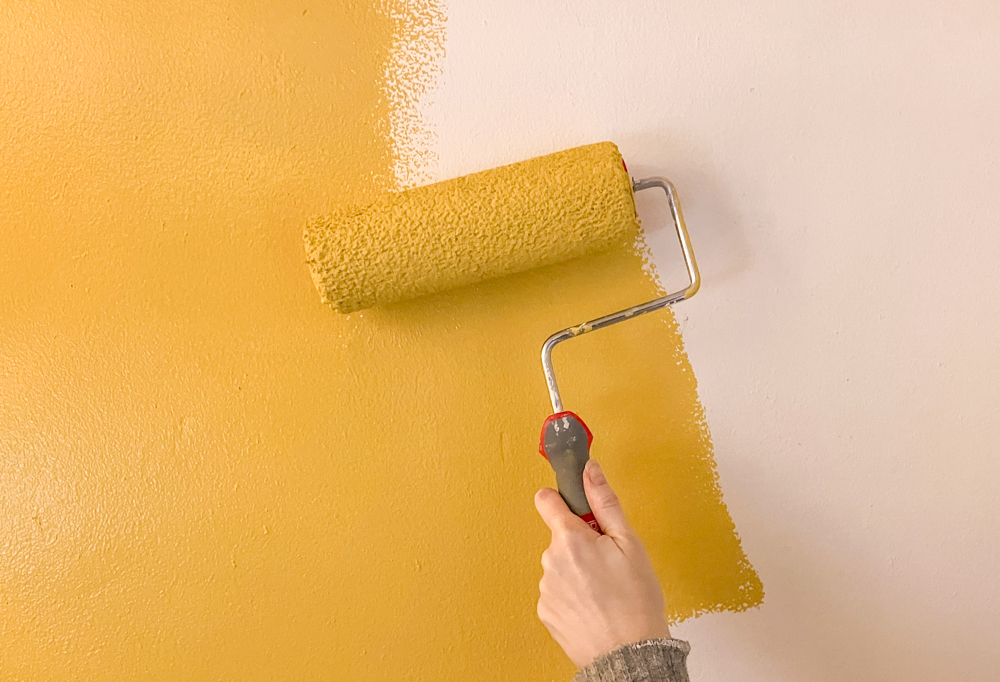 A hand holding a roller, painting a white wall with mustard yellow paint.